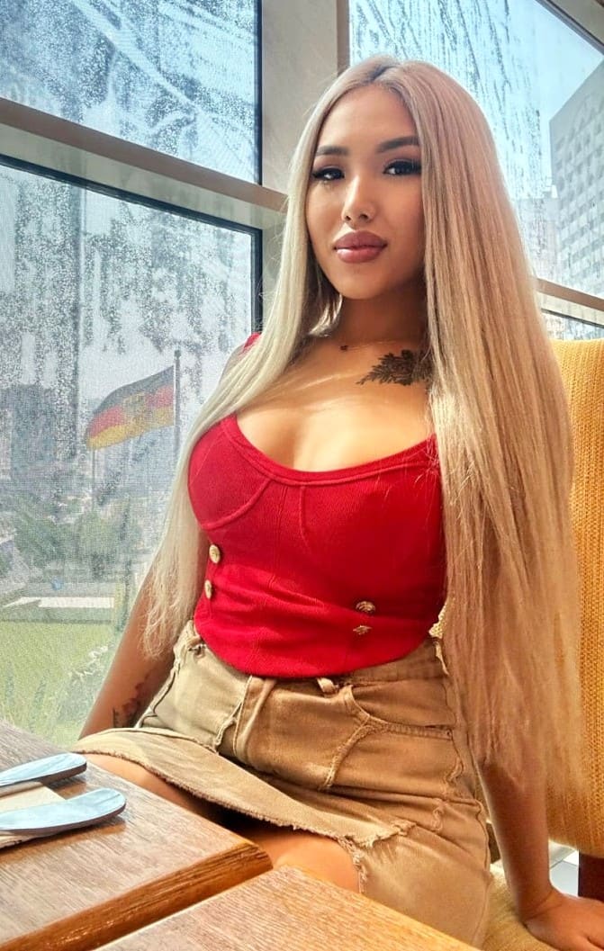 A blonde woman sits at a table by a window, wearing a red crop top and tan denim skirt with a chest tattoo visible.