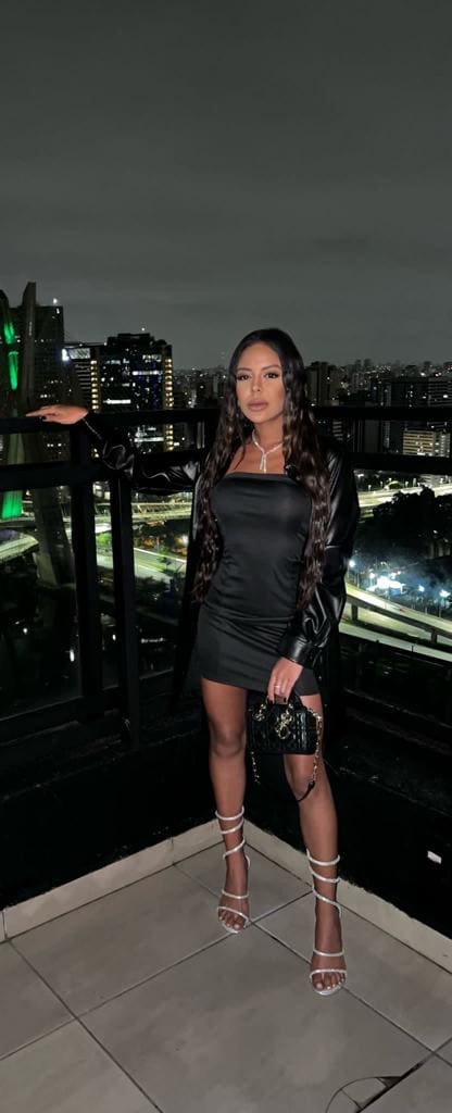 A woman in a black slip dress and leather jacket stands on a night rooftop terrace, holding a quilted bag, with city lights behind.