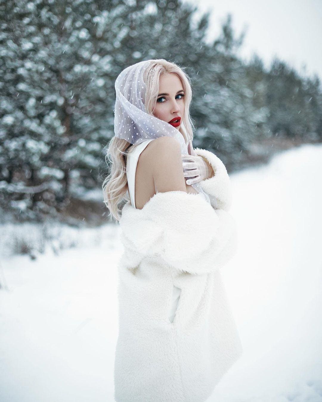 A blonde woman in a white fur coat and polka-dot scarf stands with arms crossed in a snowy landscape.