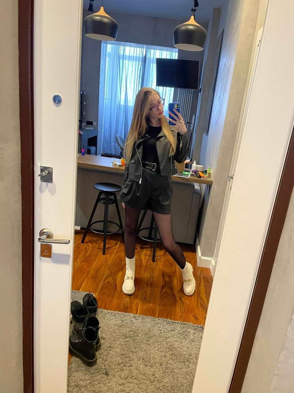 Mirror selfie in a black leather jacket, shorts, and sheer tights, with white platform shoes and boots on the floor.