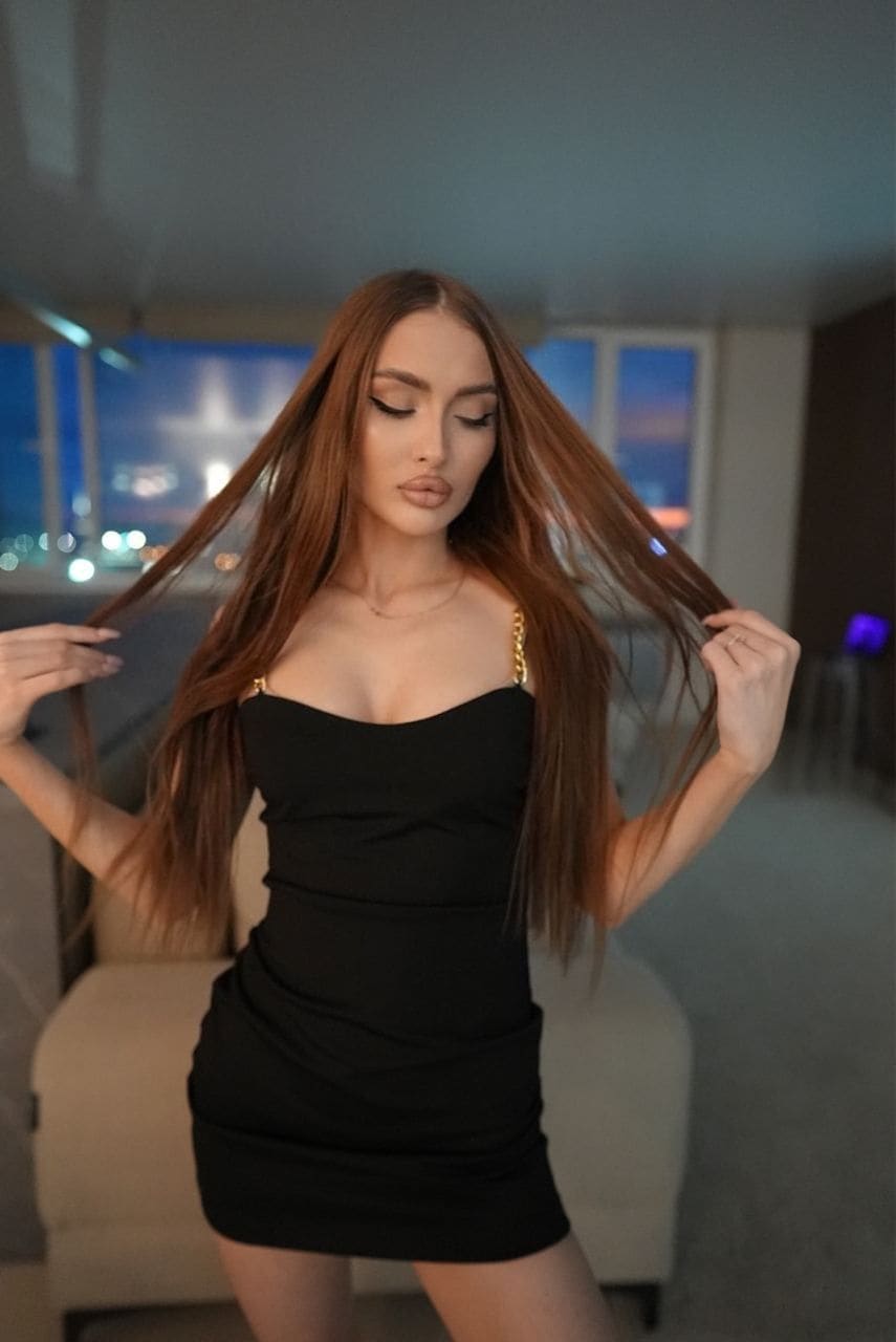 Adel stands indoors by a window with city lights, wearing a short black strapless dress with gold chain straps and holding her reddish-brown hair.