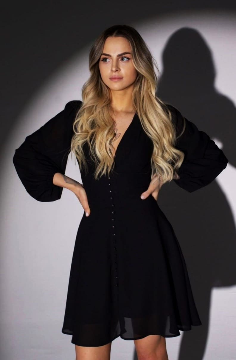 Asya stands with hands on hips in a black V-neck dress against a plain light background with a strong shadow.