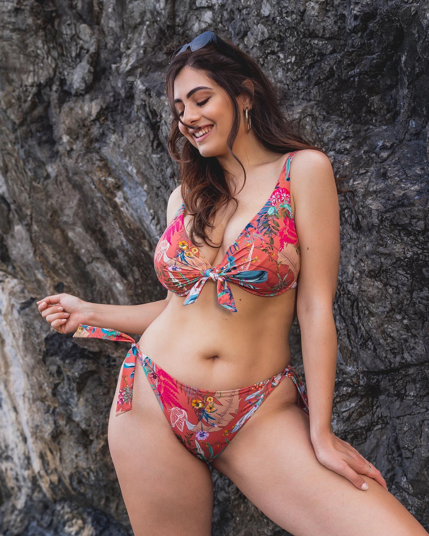 A woman in a floral bikini and sunglasses on her head stands before a dark rock wall, smiling with eyes closed.