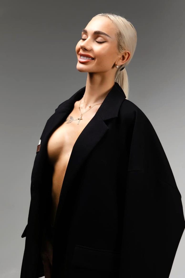 Blonde woman with eyes closed, wearing an open black blazer over a bare chest with a tattoo, against a gray studio background.