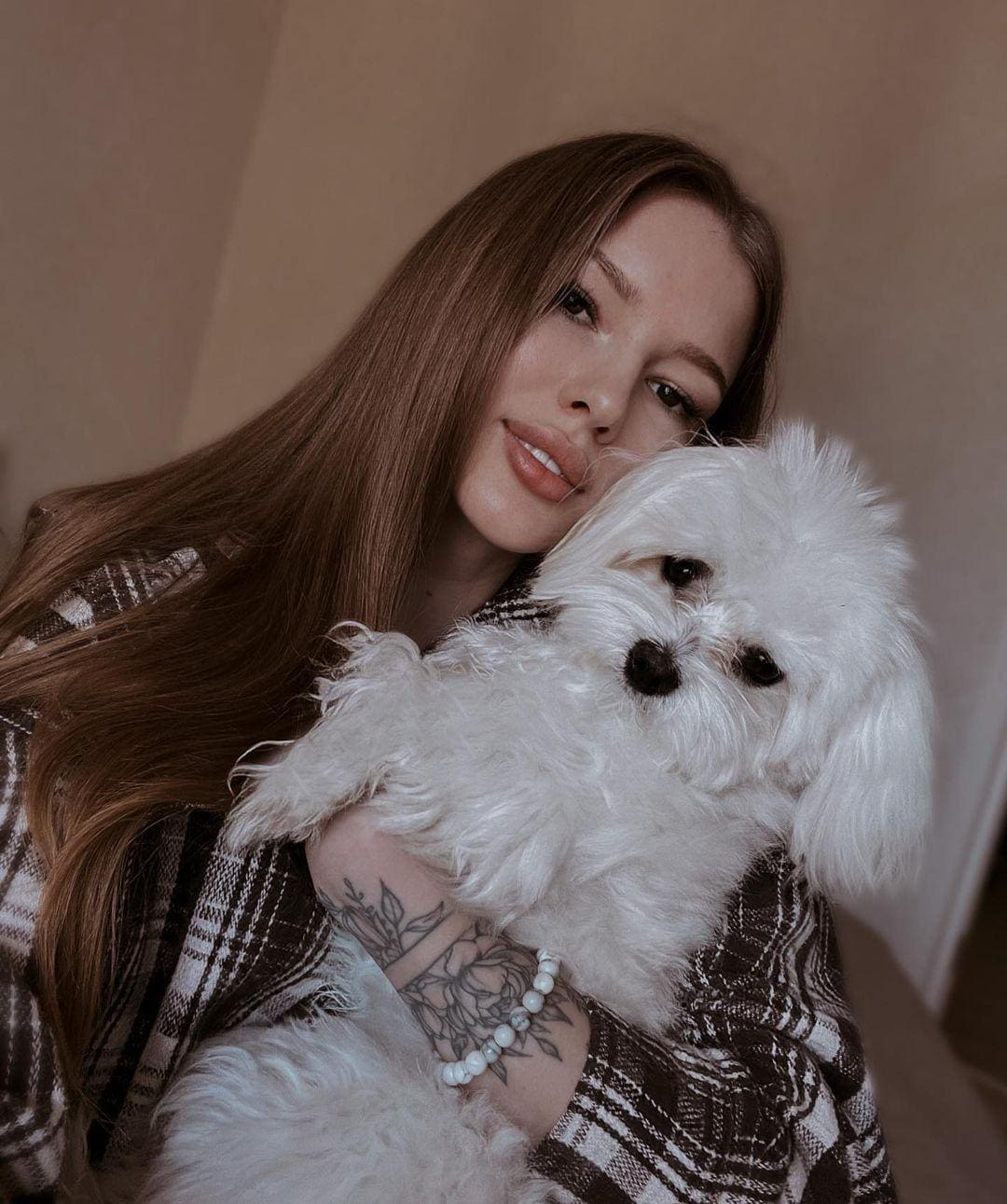Novalee holds a small white fluffy dog while wearing a brown and white plaid shirt against a plain beige wall.