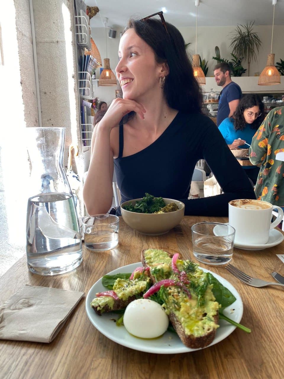 Seated at a cafe table in a black one-shoulder top, with avocado toast, egg, greens, and coffee.