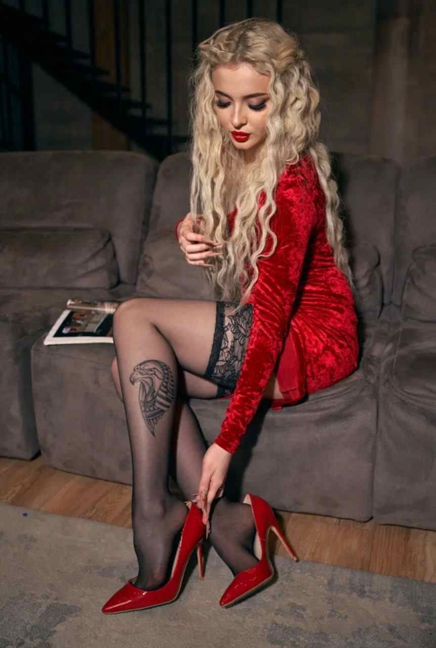 Woman in red dress and stockings sitting on a grey sofa, looking down as she adjusts her red high heel.