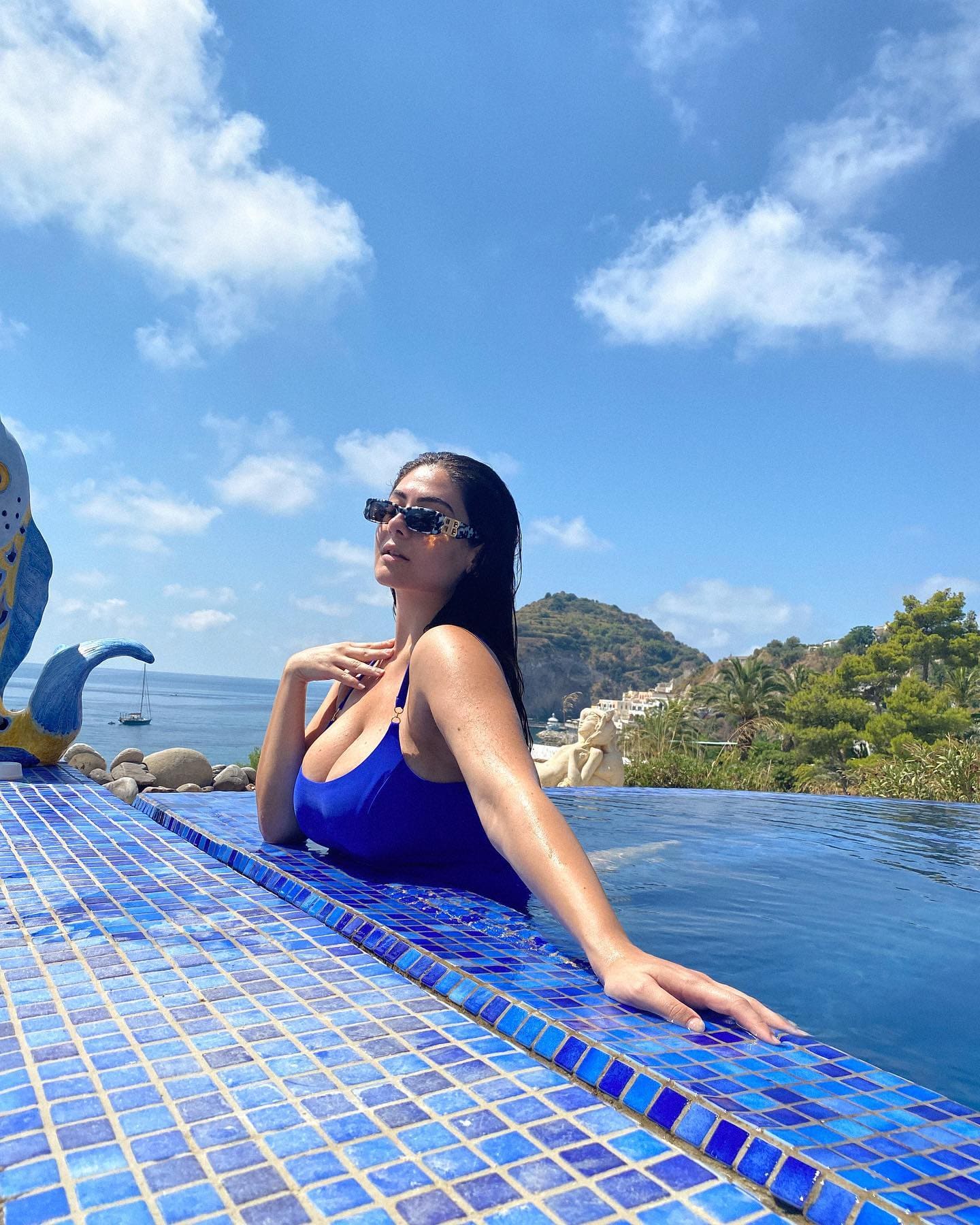A woman in a blue one-piece swimsuit and sunglasses sits at the edge of an infinity pool, coastal view behind.