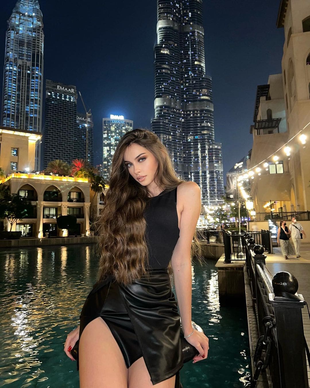 A woman stands by a canal at night in a black top and high-slit leather skirt, with a bridge and skyscrapers behind her.