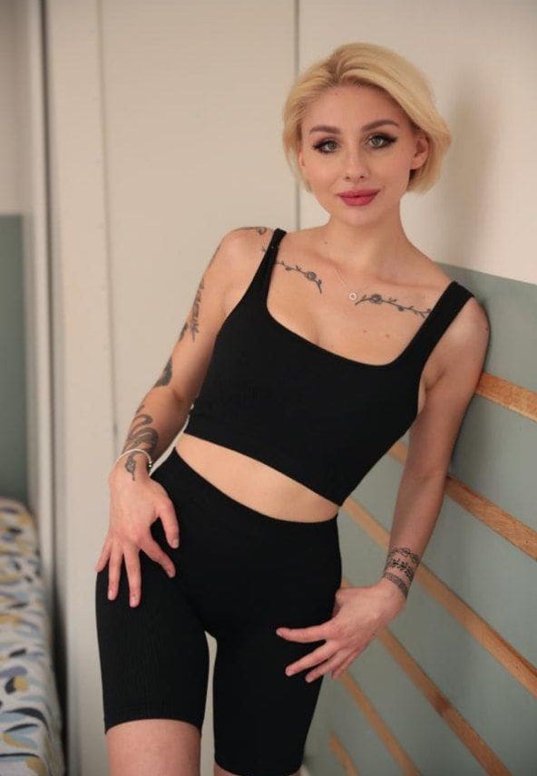 Dolores, a blonde with short hair and tattoos, leans against a wooden slatted wall wearing a black cropped tank top and mid-thigh leggings.