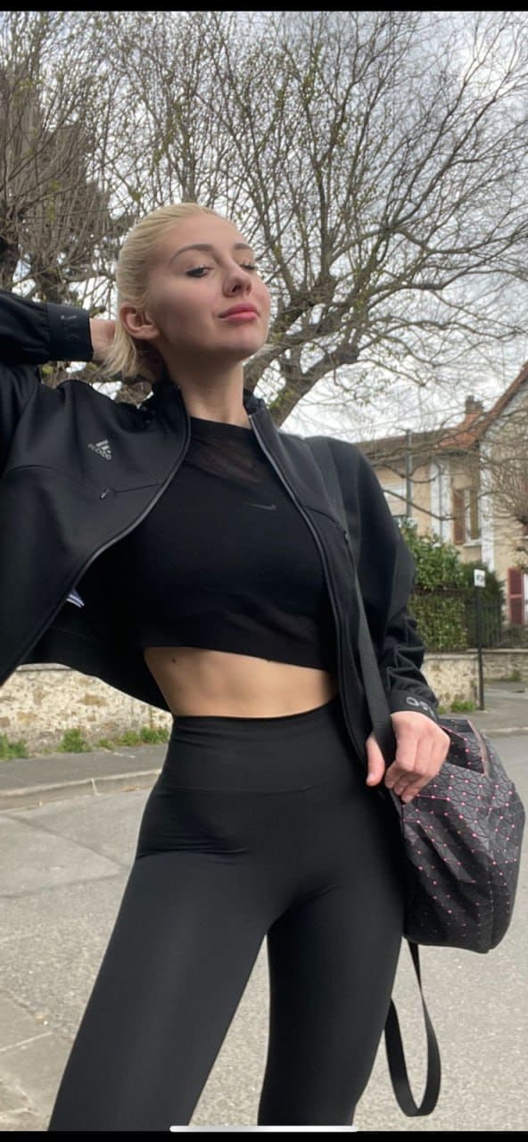 A blonde stands outdoors on a sidewalk in black athletic wear and an open jacket, holding a patterned bag with bare trees and stone in the background.