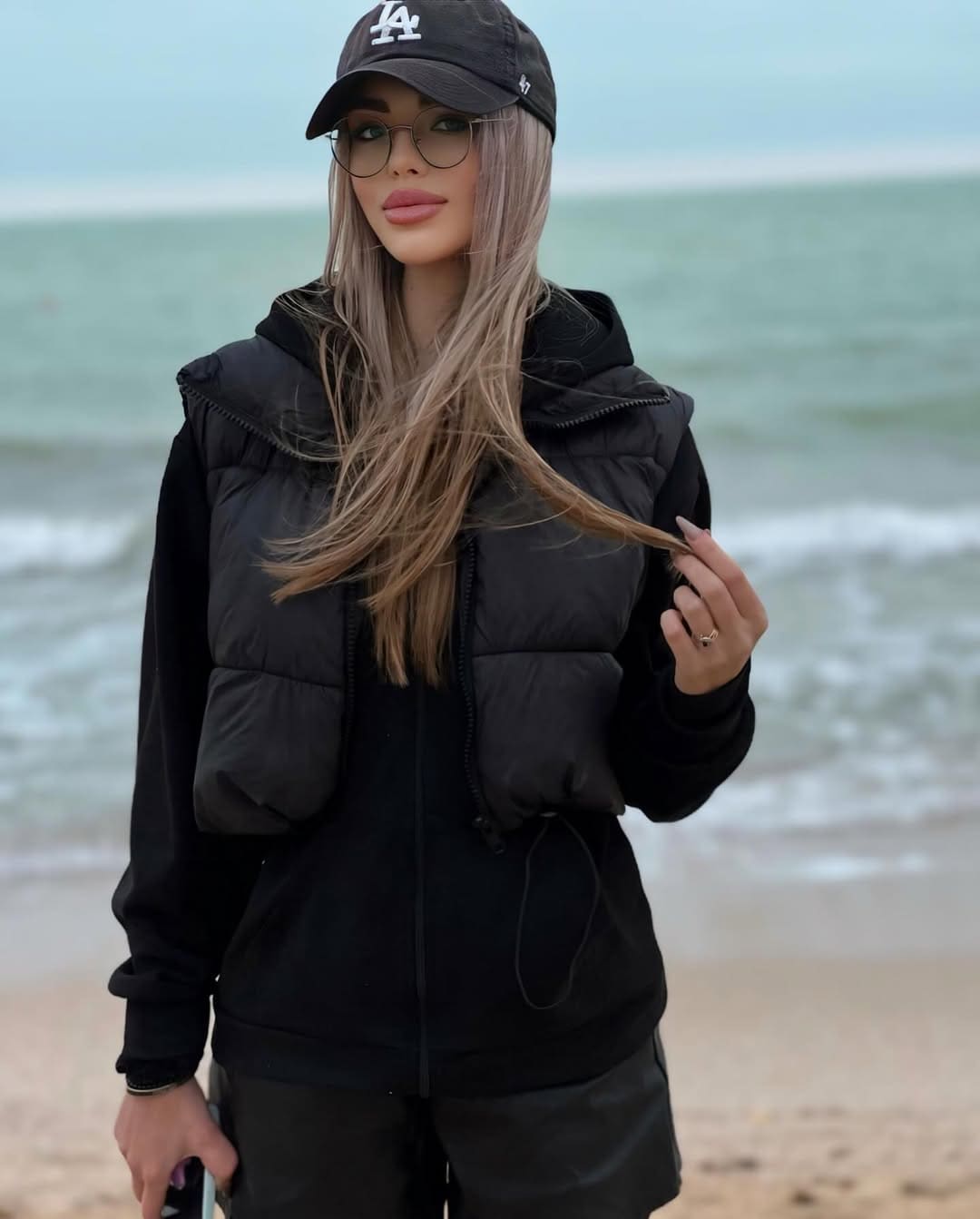 On a beach with the ocean behind, a woman in a black cap, glasses, hoodie, and puffer vest holds a strand of her blonde hair.