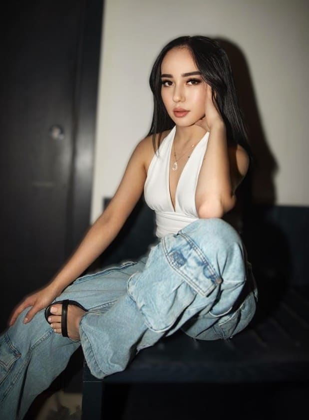 Malika sits on a dark surface in a white halter top and baggy jeans, one hand behind her head, against a plain wall and dark door.