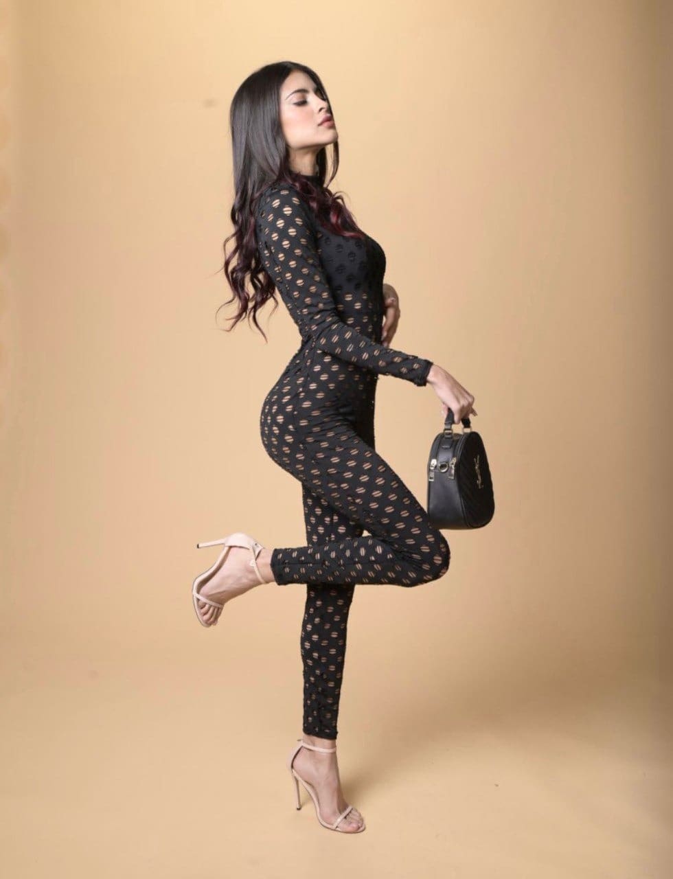 Standing in profile against a tan backdrop, she wears a dotted black jumpsuit and nude heels, lifting one leg behind her.
