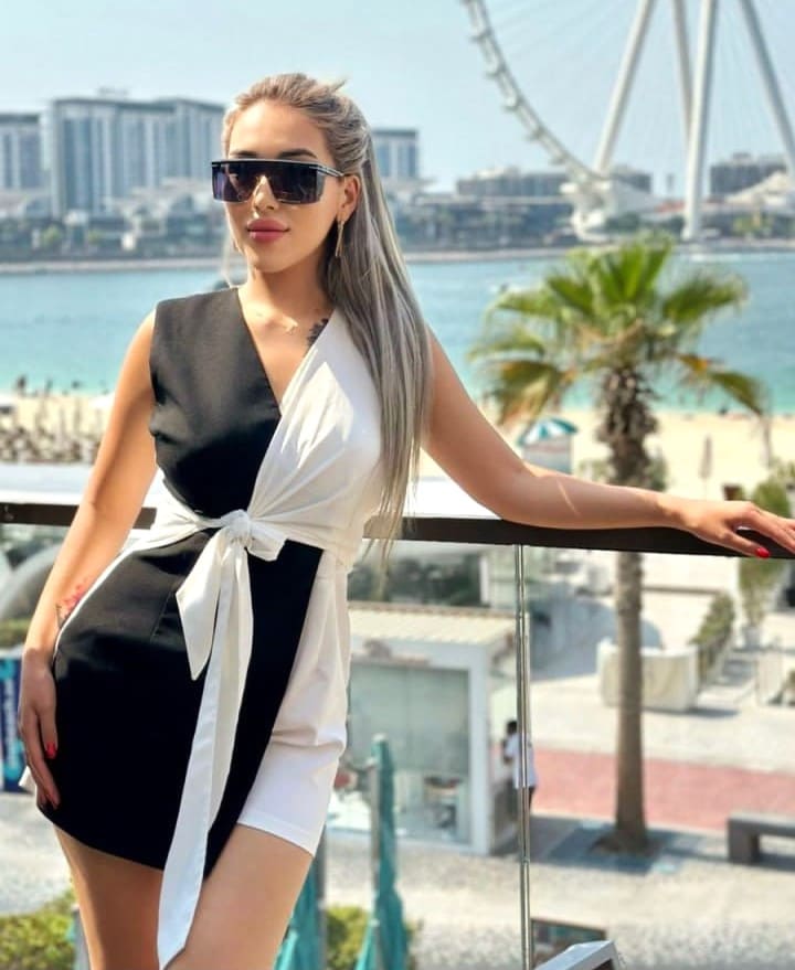 A woman in sunglasses and a black-and-white wrap dress stands on a balcony overlooking a city and Ferris wheel.