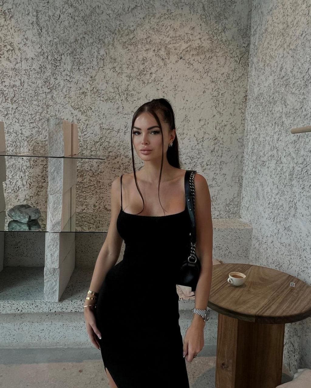 Eleanor stands by stone walls in a black sleeveless dress, holding a chain-strap bag near a small wooden table.