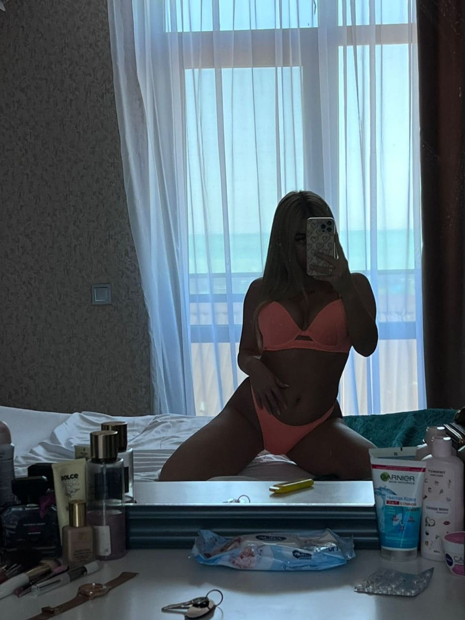 A woman kneels on a bed taking a mirror selfie in a coral bikini, with a vanity and window in the background.